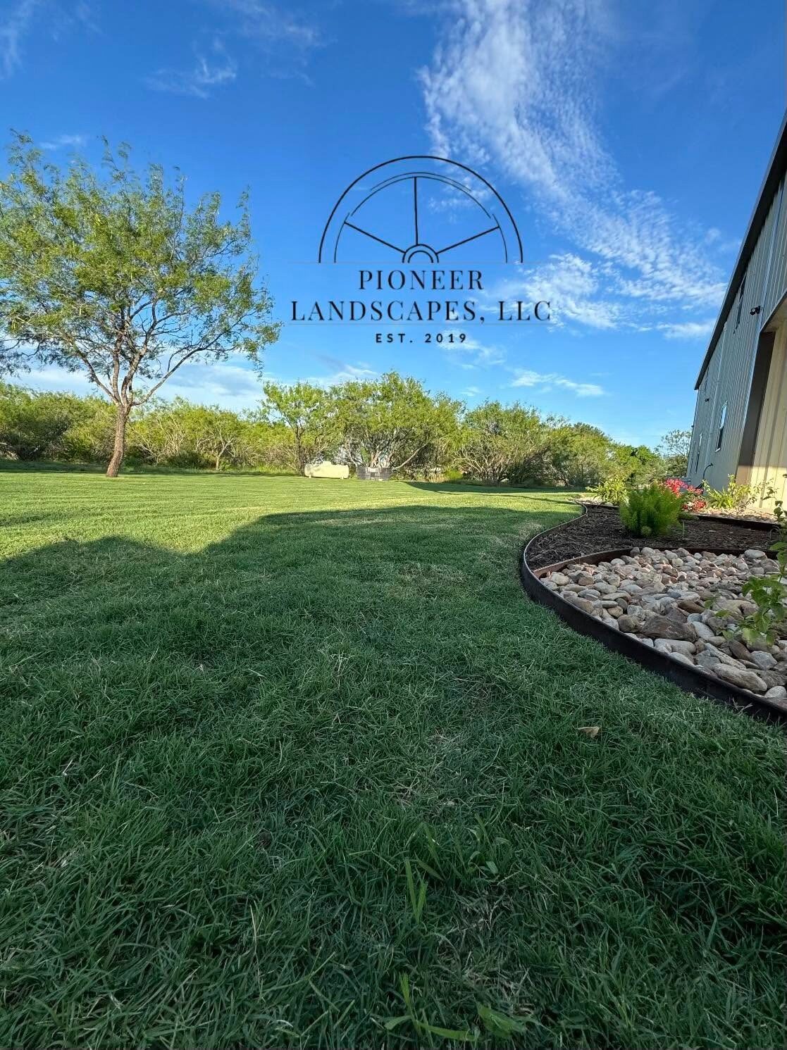 A finished Pioneer install: established lawn with a steel-edged river rock bed and plantings on a country property