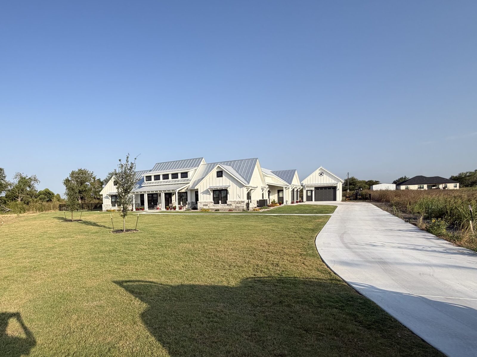 Wide view of the finished property: established lawn across a large open lot with young trees, a long concrete drive, and open country beyond