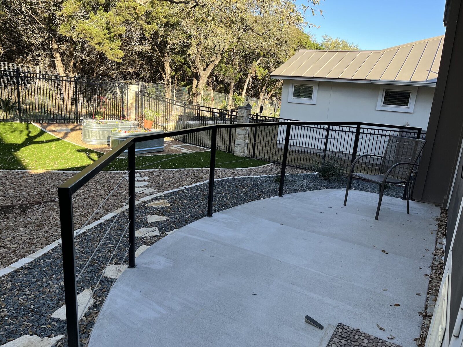 The railing curving along the patio with the back yard, gravel beds, and stock tank planters beyond
