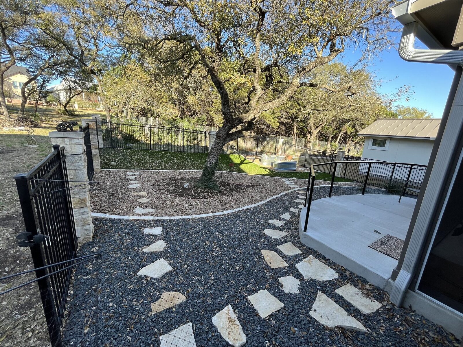 A finished Pioneer back yard: flagstone stepping paths through dark gravel, a limestone-edged tree well around a mature live oak, and a poured patio with black steel railing