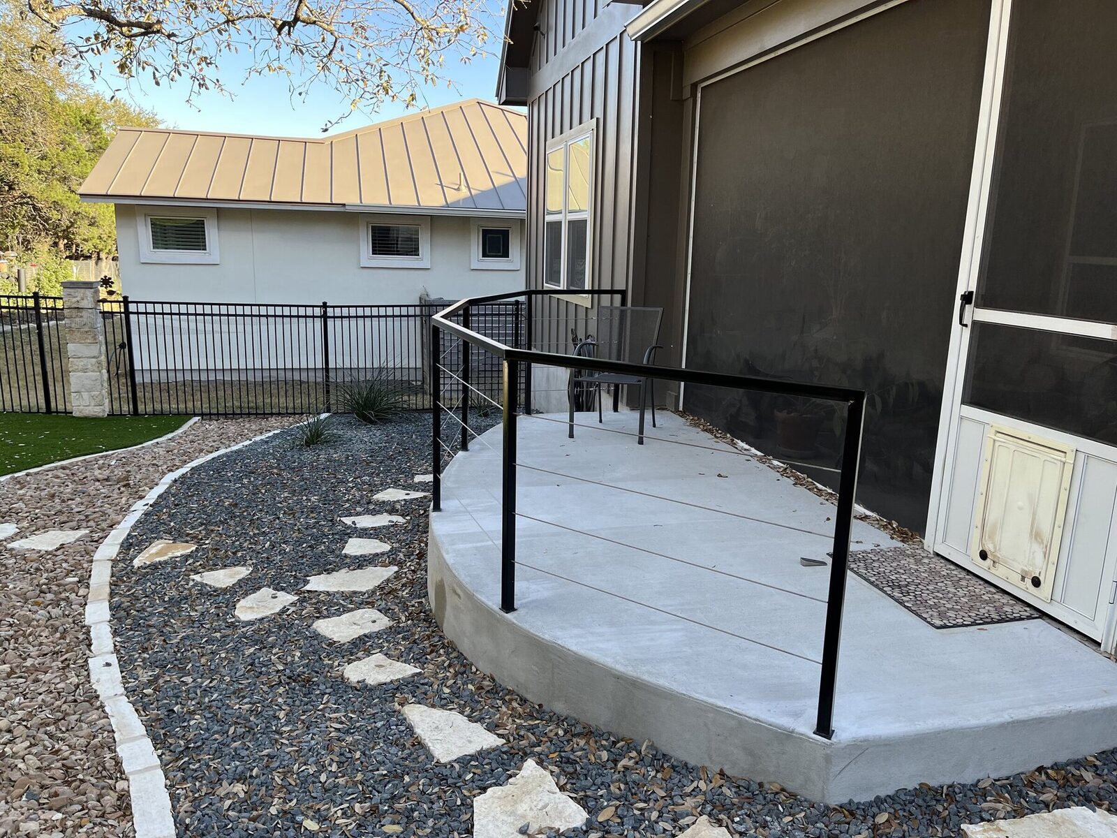 The curved concrete patio with its custom black steel cable railing, flagstone steppers, and gravel beyond
