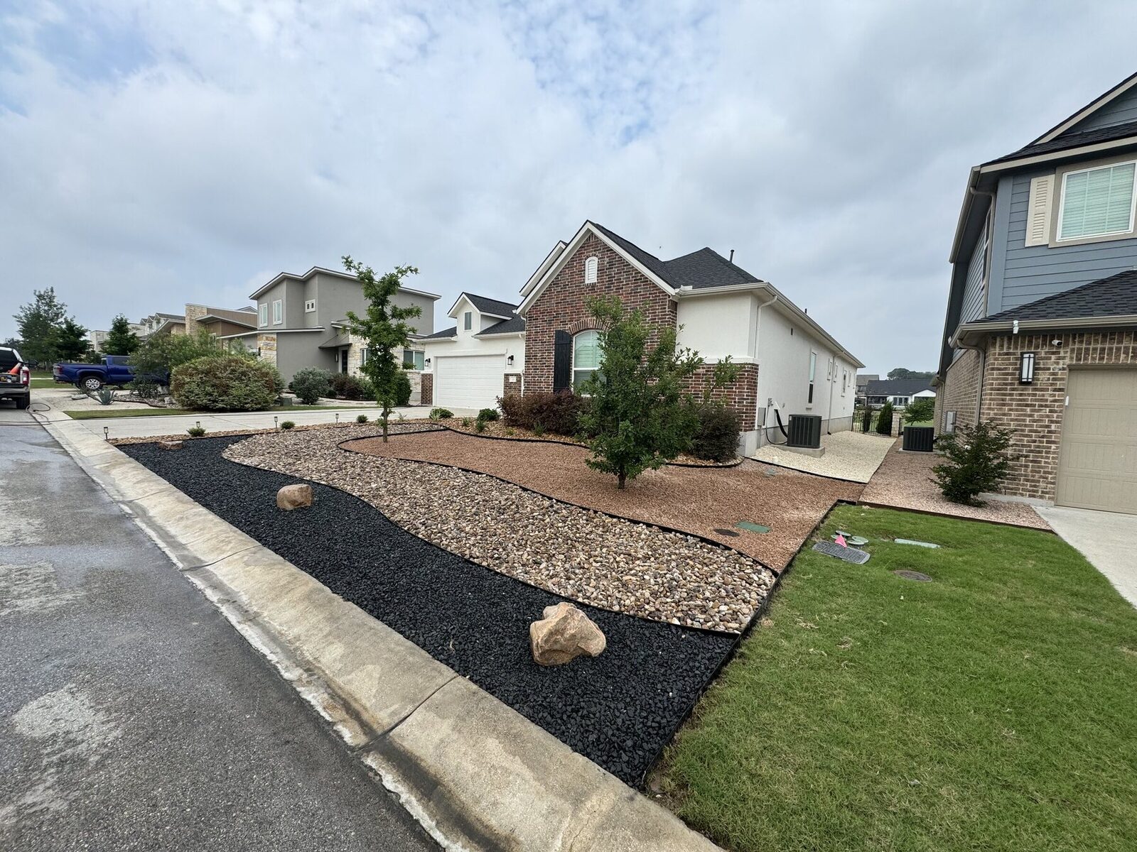 A finished Pioneer xeriscape: curved bands of black basalt and tan river rock with feature boulders and retained trees across a front yard