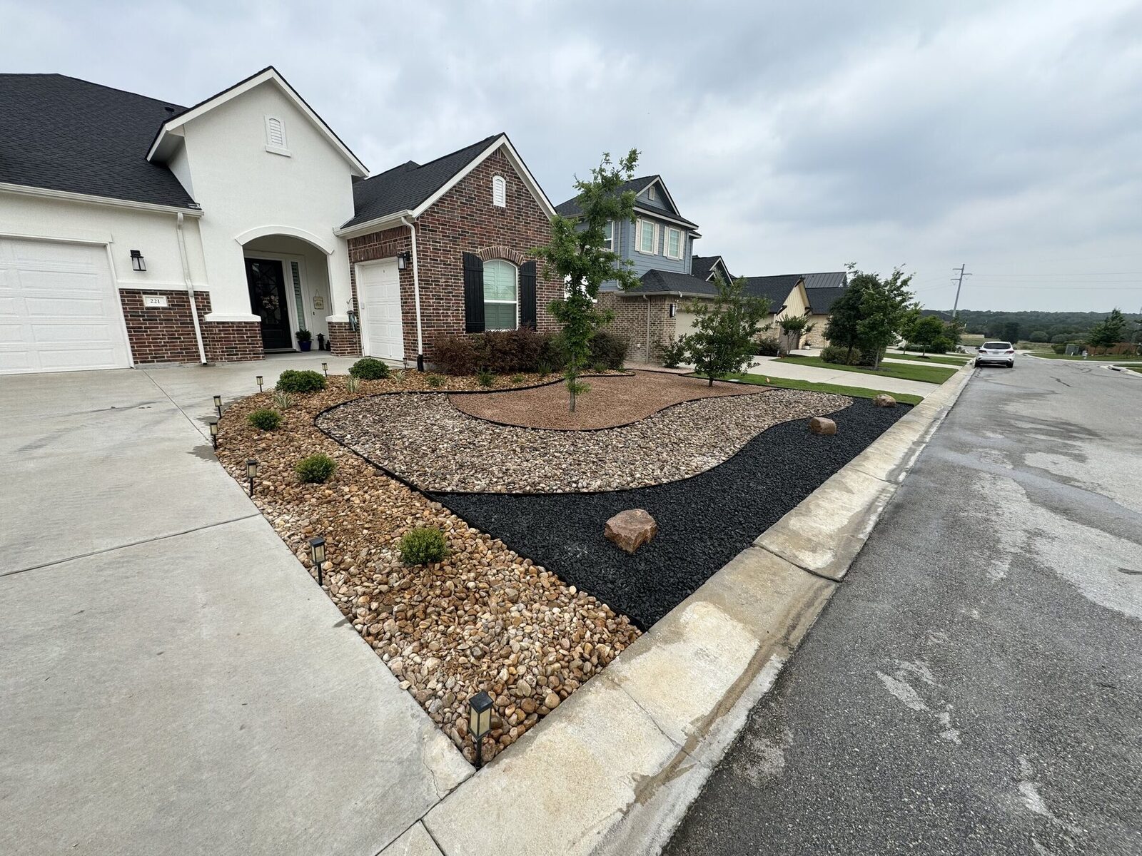 The finished xeriscape from the corner: three stone zones in curved bands with a boulder, path lights, and a young shade tree